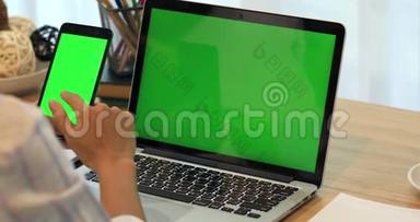 亚洲女人手<strong>拿手机</strong>。 带有绿色屏幕的桌子上的电话和笔记本电脑。